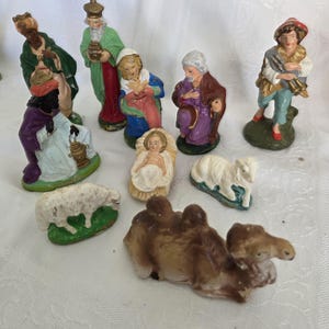 May include: A collection of ten hand-painted ceramic nativity figurines including figures of the holy family, three wise men, a shepherd, two sheep, and a camel. Each figure is depicted in colorful robes and stands on a small green base.