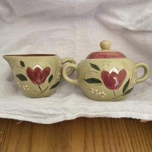 May include: A cream and burgundy floral creamer and sugar bowl set. The set features a light green base color with burgundy tulip designs, green leaves, and small white flower accents. The sugar bowl has a matching lid.