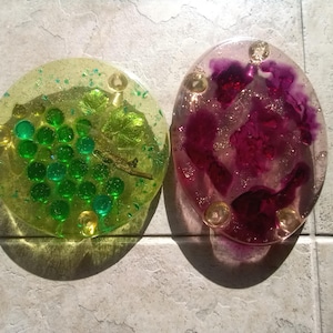 May include: Two round, clear resin coasters with embedded objects. One coaster has green and gold glitter with green beads and a dried leaf. The other coaster has purple and gold glitter with a dried flower.