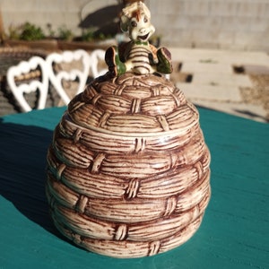 May include: A ceramic honey pot shaped like a beehive with a cartoon bee figurine on top. The beehive is brown and white with a textured surface. The bee is yellow, black, and green.