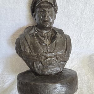 May include: A bronze-colored bust sculpture of a man wearing a military cap and uniform. The sculpture is mounted on a circular base. The bust depicts the man's head, shoulders, and upper chest.