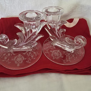 May include: Two clear glass candle holders with ornate floral designs. The candle holders have a decorative base and a detailed, swirling design. They are resting on a red cloth.