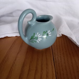 May include: A small, light blue ceramic pitcher with a white floral design. The pitcher has a rounded body and a curved handle.