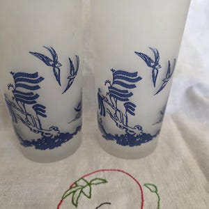 May include: Two frosted glass tumblers with a blue bird and landscape design. The glasses have a cylindrical shape and are decorated with a blue illustration of birds and a landscape scene. The glasses are sitting on a white surface.