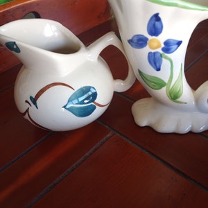 May include: Two ceramic pitchers with painted floral designs. One pitcher is white with blue and green flowers, the other is white with brown and blue leaves.