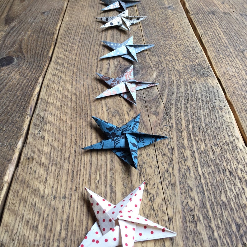 May include: A row of origami stars in various patterns and colors, including blue floral, white with red polka dots, and patterned designs. The stars are arranged on a rustic wooden surface, creating a decorative display.
