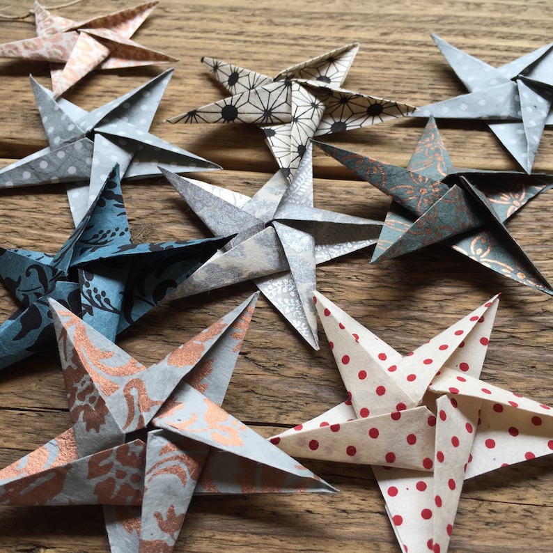 May include: A collection of handmade origami stars in various colors and patterns. The stars are made from patterned paper, including polka dots, floral designs, and geometric shapes. The color palette includes shades of blue, gray, red, and copper.