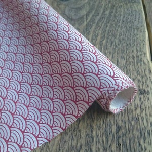 May include: A piece of fabric featuring a repeating pattern of white, concentric semicircles arranged in a fan-like design against a solid red background. The fabric is rolled at one end, revealing a white underside with a finished stitched edge.