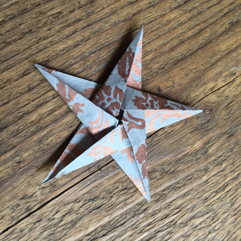 May include: A handmade origami star with five points. The star is light blue with a copper-colored floral pattern. The star is displayed on a rustic wooden surface.