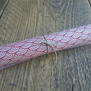 May include: A cylindrical roll of paper featuring a repeating pattern of white scalloped arches on a deep red background, secured in the center with a thin piece of natural brown twine tied into a small bow.