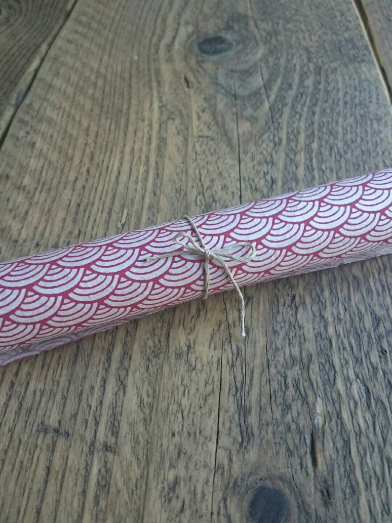 May include: A cylindrical roll of paper featuring a repeating pattern of white scalloped arches on a deep red background, secured in the center with a thin piece of natural brown twine tied into a small bow.