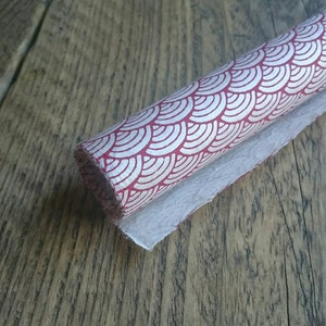 May include: A roll of textured paper featuring a repeating red and white fan-shaped scale pattern. The paper has a fibrous, handmade appearance with a visible grain and a slightly uneven edge along the length of the roll, showing a white underside.