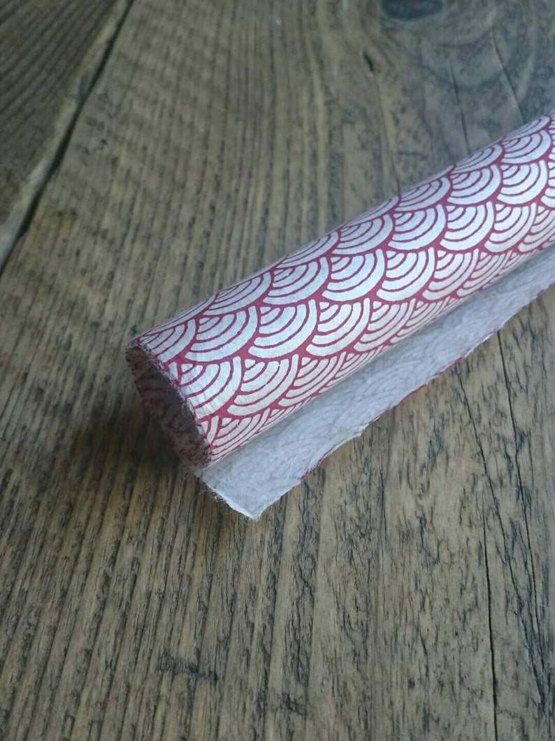 May include: A roll of textured paper featuring a repeating red and white fan-shaped scale pattern. The paper has a fibrous, handmade appearance with a visible grain and a slightly uneven edge along the length of the roll, showing a white underside.