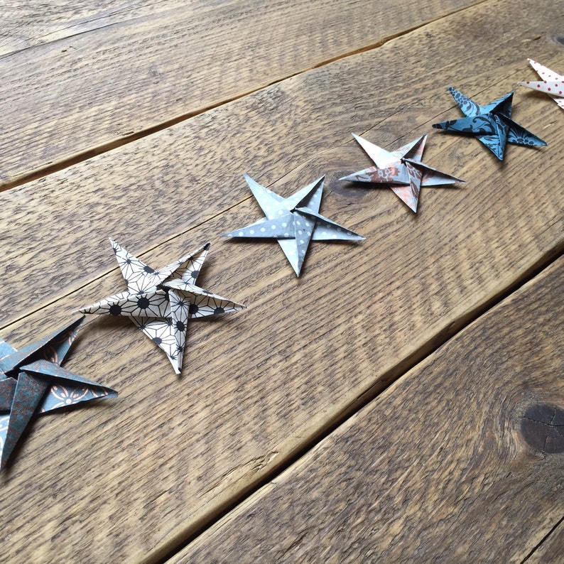 May include: A line of origami stars in various patterns and colors, including blue, white, and patterned paper, are arranged diagonally on a rustic wooden surface. The stars are folded with intricate detail, showcasing a handmade craft.