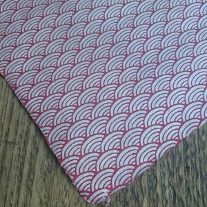 May include: A piece of fabric featuring a repeating pattern of white, concentric semicircles arranged in rows against a solid red background. The material has a visible texture and a slightly frayed edge, displaying a consistent geometric design.