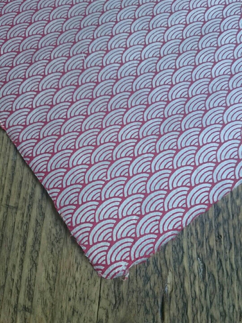 May include: A piece of fabric featuring a repeating pattern of white, concentric semicircles arranged in rows against a solid red background. The material has a visible texture and a slightly frayed edge, displaying a consistent geometric design.