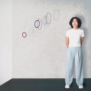 May include: A woman with curly brown hair stands in front of a white wall with a mobile hanging from the ceiling. The mobile has red and blue circles and is made of thin metal.