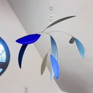 May include: A modern mobile sculpture with a minimalist design. It features curved metal rods suspending leaf-shaped elements in shades of blue, gray, and silver. The mobile hangs from a thin wire, creating a sense of movement and balance.