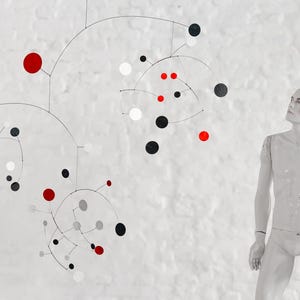 May include: A mobile sculpture with black, white, and red circles hanging from thin wires. The sculpture is suspended from the ceiling and is in motion. A white mannequin is standing to the right of the sculpture.