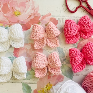 May include: Four handmade crocheted hair bows in pink and white. The bows are arranged on a floral background.