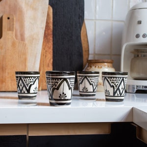 Puede incluir: Cuatro tazas de cerámica con diseños en blanco y negro se colocan sobre una encimera blanca. Las tazas tienen un diseño geométrico. Detrás de las tazas hay tablas de cortar de madera y una cafetera blanca.