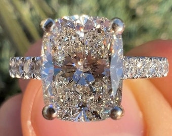 Könnte beinhalten: Ein Kissen-Schliff-Diamant-Verlobungsring mit einer Pavé-Fassung. Der Ring ist in Weißgold gefasst und verfügt über einen großen, funkelnden Diamanten.