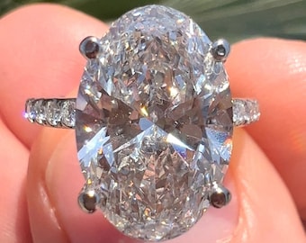 May include: A large, oval-shaped diamond engagement ring set in a white gold band. The diamond is surrounded by smaller diamonds on the band.