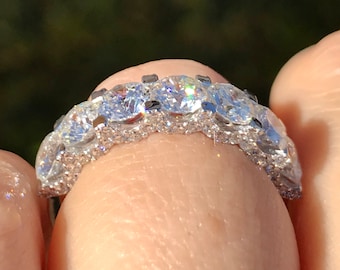 May include: A close-up of a silver ring with multiple clear, round-cut stones. The ring is set with a row of larger stones and smaller stones along the band. The ring is held between two fingers.