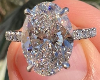 Könnte beinhalten: Ein silberner Ring mit einem großen ovalen Diamanten, der in einer Krappenfassung sitzt. Der Diamant ist von kleineren Diamanten umgeben, die einen Heiligenschein um ihn bilden. Die Ringschiene ist ebenfalls mit kleineren Diamanten besetzt.