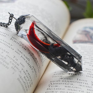 May include: A clear resin pendant with a black chain. The pendant has a rectangular shape with a red curved element and black inclusions. The pendant is resting on an open book.