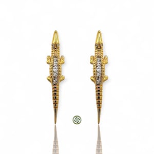 May include: A pair of gold-colored alligator earrings. Each earring features a detailed alligator design with a textured body and sparkling accents. The earrings are displayed on a white background with a reflection.
