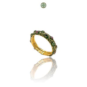Peut inclure: Un jonc en or avec un motif en émail vert et des pierres précieuses incrustées. Le jonc présente un motif répétitif de volutes ornées et est orné de pierres rouges et vert foncé. Le bijou est présenté sur fond blanc avec son reflet visible.