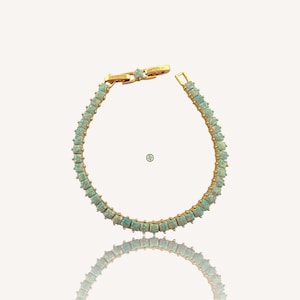 Puede incluir: Una pulsera dorada con una serie de piedras preciosas cuadradas de color verde pálido. La pulsera tiene una cadena delicada y un cierre seguro. Las piedras están espaciadas uniformemente, creando un patrón continuo. La pulsera se muestra sobre un fondo blanco. Las piedras miden aproximadamente 0,5 cm.