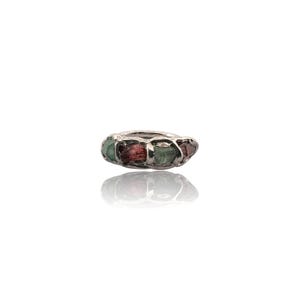 May include: A silver ring with a row of rough-cut gemstones in red and green hues. The ring's design highlights the natural, irregular shapes of the stones, set in a textured silver band. The ring is displayed on a white surface.