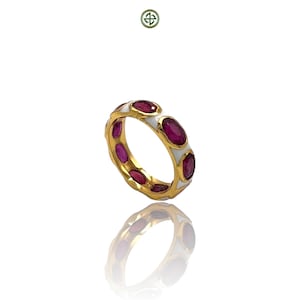 Puede incluir: Un anillo de oro con piedras preciosas ovaladas de color rojo intenso engastadas en una banda de esmalte blanco. El diseño del anillo presenta un patrón repetitivo de piedras preciosas y esmalte, creando un aspecto continuo y elegante. El anillo se muestra sobre una superficie reflectante.