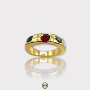 May include: A gold band ring with three gemstones. The center stone is a red ruby, and the two side stones are green. The ring has a textured surface.