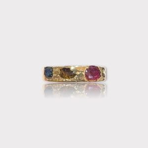 May include: A gold band ring with three colorful gemstones. The ring features a blue sapphire, a brown and yellow gemstone, and a red ruby.
