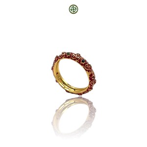 Könnte beinhalten: Ein goldener Ring mit rotem Emaille-Design und kleinen grünen Akzenten. Der Ring weist ein verziertes Muster auf und wird auf weißem Hintergrund mit seiner darunter sichtbaren Reflexion dargestellt.