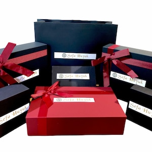 Peut inclure: Une collection de six bo&icirc;tes cadeaux, toutes noires avec des accents rouges. Les bo&icirc;tes sont attach&eacute;es avec un ruban rouge et portent une &eacute;tiquette dor&eacute;e avec le texte "Sefa Huyuk". Les bo&icirc;tes sont dispos&eacute;es en forme de pyramide, la plus grande en bas et la plus petite en haut.