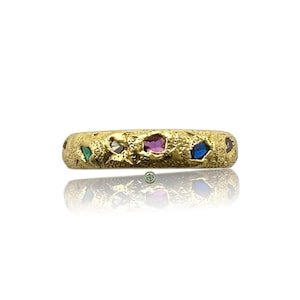 Peut inclure: Une bague en or avec une surface texturée, sertie de diverses pierres précieuses colorées. La bague présente un mélange de pierres vertes, violettes, bleues et claires, créant un design unique et accrocheur. La bague est présentée sur fond blanc.