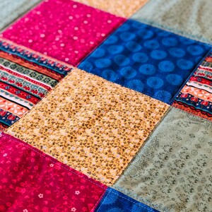 Op de afbeelding: Een patchworkdeken met vierkanten in verschillende kleuren en patronen, waaronder roze, blauw, geel en groen.