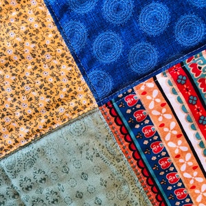 Op de afbeelding: Een patchworkdeken met vier verschillende vierkanten van stof. De linkerbovenste vierkant is een gele stof met een klein wit bloemenpatroon. De rechterbovenste vierkant is een blauwe stof met een wit cirkelpatroon. De linkeronderste vierkant is een groene stof met een klein wit bloemenpatroon. De rechteronderste vierkant is een veelkleurige stof met een gestreept patroon.