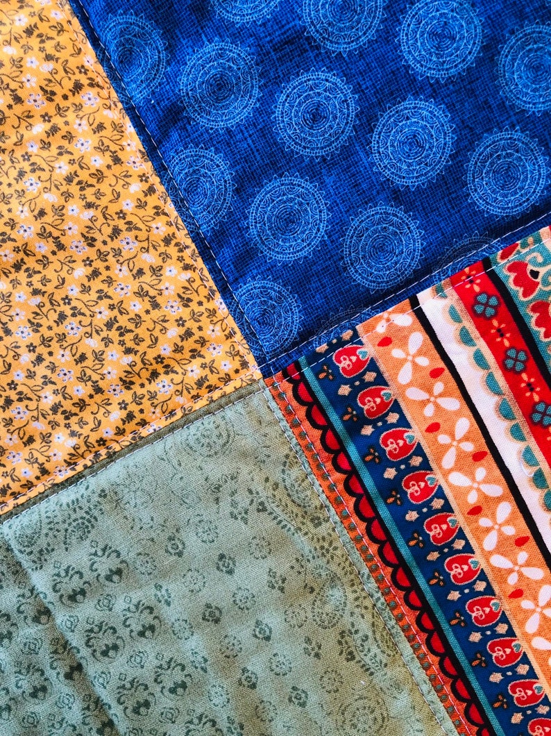Op de afbeelding: Een patchworkdeken met vier verschillende vierkanten van stof. De linkerbovenste vierkant is een gele stof met een klein wit bloemenpatroon. De rechterbovenste vierkant is een blauwe stof met een wit cirkelpatroon. De linkeronderste vierkant is een groene stof met een klein wit bloemenpatroon. De rechteronderste vierkant is een veelkleurige stof met een gestreept patroon.