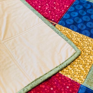 Op de afbeelding: Een close-up van een quilt met een patchwork-ontwerp. De quilt is gemaakt van verschillende stoffen in tinten groen, rood, geel en blauw. De quilt is gestikt met een eenvoudig steekpatroon. De quilt is over zichzelf gevouwen, waardoor de achterkant zichtbaar wordt, die een effen beige kleur heeft.