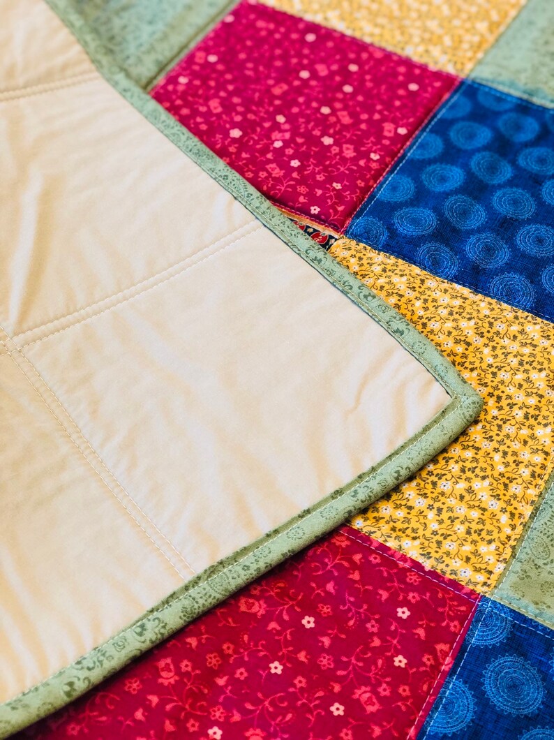 Op de afbeelding: Een close-up van een quilt met een patchwork-ontwerp. De quilt is gemaakt van verschillende stoffen in tinten groen, rood, geel en blauw. De quilt is gestikt met een eenvoudig steekpatroon. De quilt is over zichzelf gevouwen, waardoor de achterkant zichtbaar wordt, die een effen beige kleur heeft.