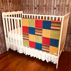 Op de afbeelding: Een wit wiegje met een wit gerimpeld wiegje rok en een kleurrijk patchwork dekbed. Het dekbed heeft vierkanten van rode, blauwe, gele en groene stof.