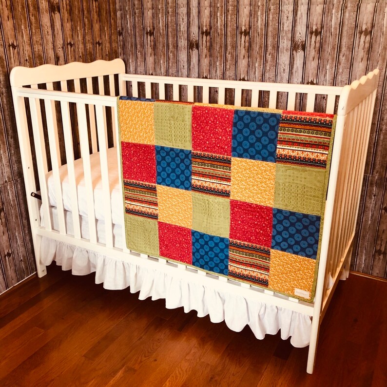 Op de afbeelding: Een wit wiegje met een wit gerimpeld wiegje rok en een kleurrijk patchwork dekbed. Het dekbed heeft vierkanten van rode, blauwe, gele en groene stof.