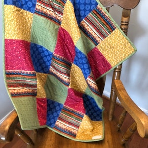 Op de afbeelding: Een patchworkdeken met vierkanten van rode, blauwe, groene en gele stof. De deken is gemaakt van verschillende patronen, waaronder stippen, strepen en bloemenpatronen. De deken is over een houten schommelstoel gelegd.