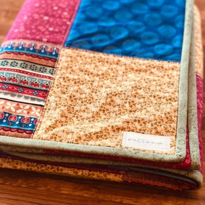 Op de afbeelding: Een veelkleurig patchwork-deken met een groene rand en een wit label met de tekst "putt-o-w".