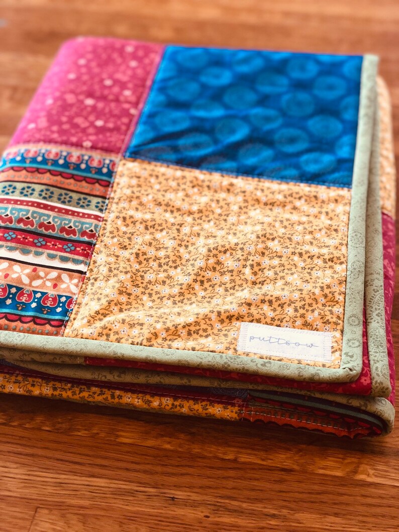 Op de afbeelding: Een veelkleurig patchwork-deken met een groene rand en een wit label met de tekst "putt-o-w".